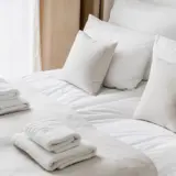 Cropped view of white bedclothes and towels on bed in comfortable hotel room, hypoallergenic pillows, personal comfort idea, bedding concept, bathroom details